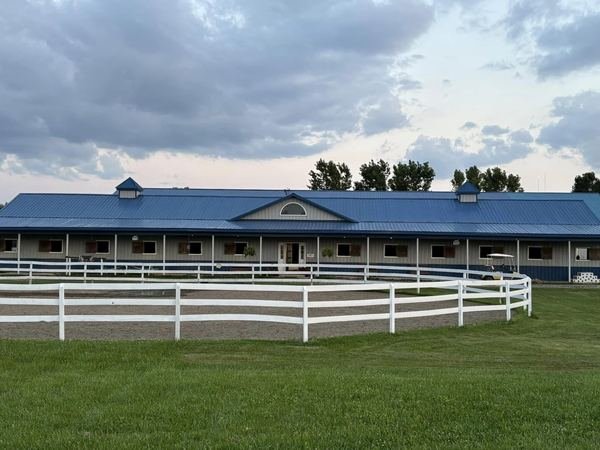 The main barn at Swan Creek Stables, a long single-storey stable with a blue metal roof and covered walkway, seen across the white-fenced outdoor arena