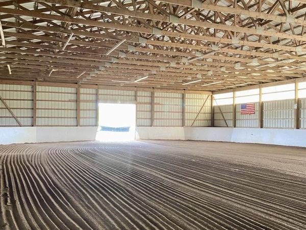 The indoor riding arena with freshly dragged sand footing, open timber roof trusses and a wide door letting in daylight