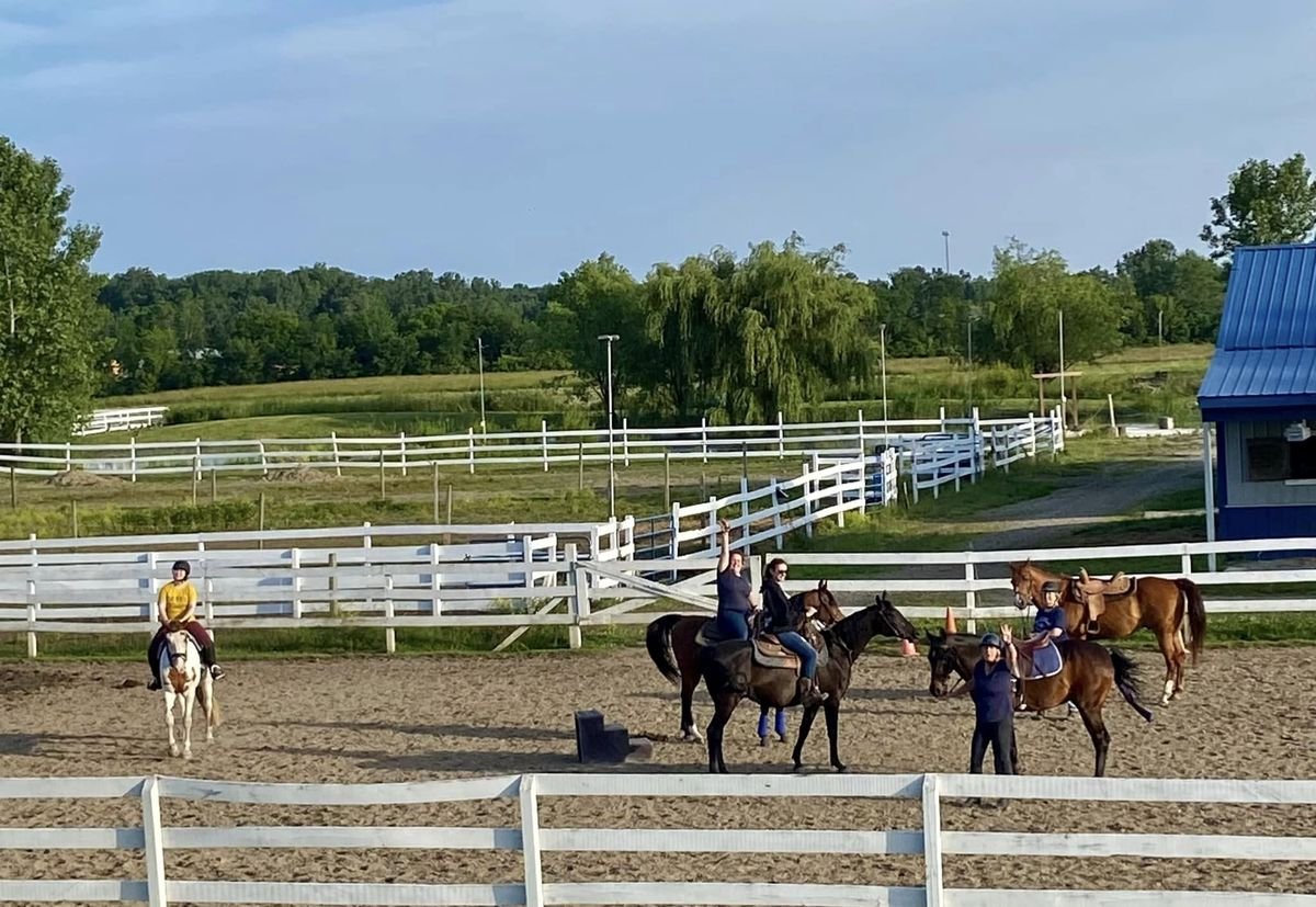 Riders and horses gathered in the sand outdoor arena on a summer evening, with white fencing and green pasture behind them