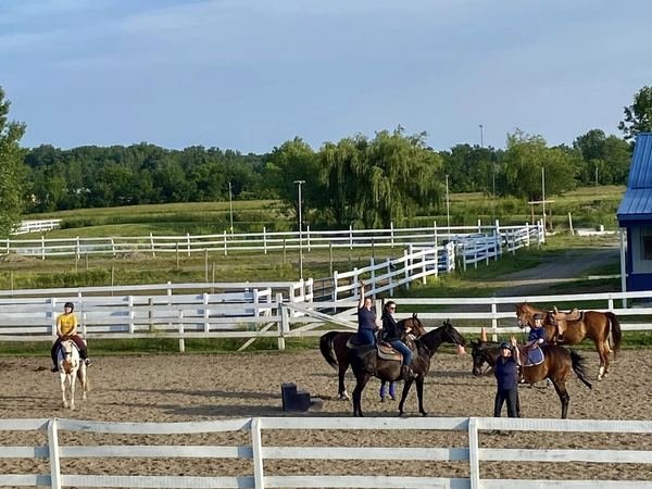 Riders and horses gathered in the sand outdoor arena on a summer evening, with white fencing and green pasture behind them