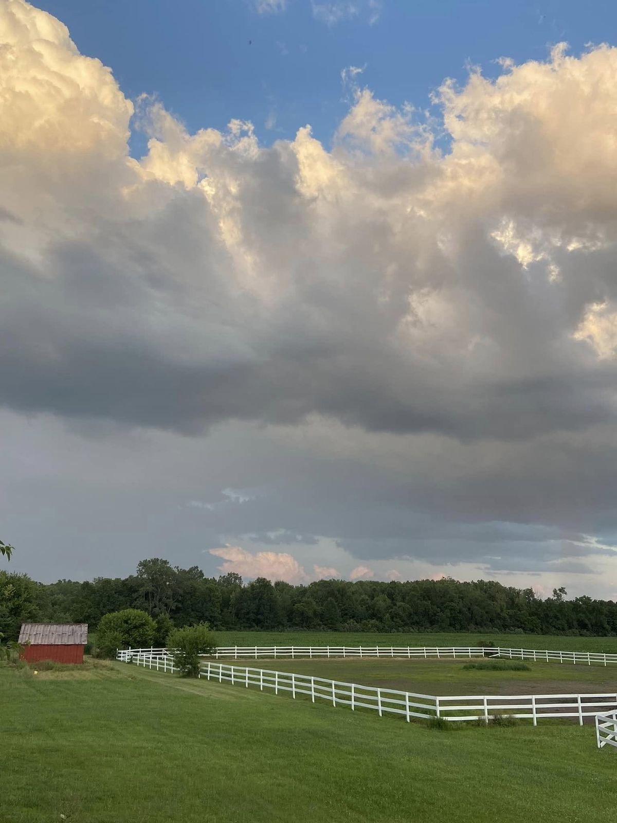 Wide green pasture bordered by white three-rail fencing under tall summer clouds, with a small red barn at the edge of the treeline